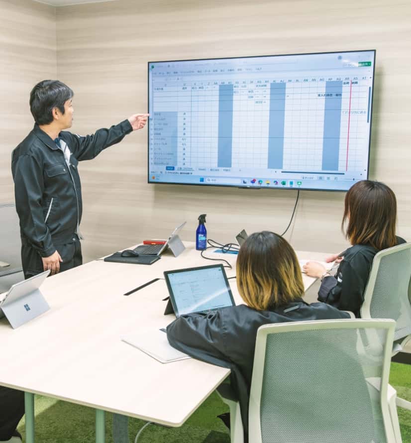 画面の資料を指さしながら説明をする男性社員と、座って話を聞いている社員２名の写真