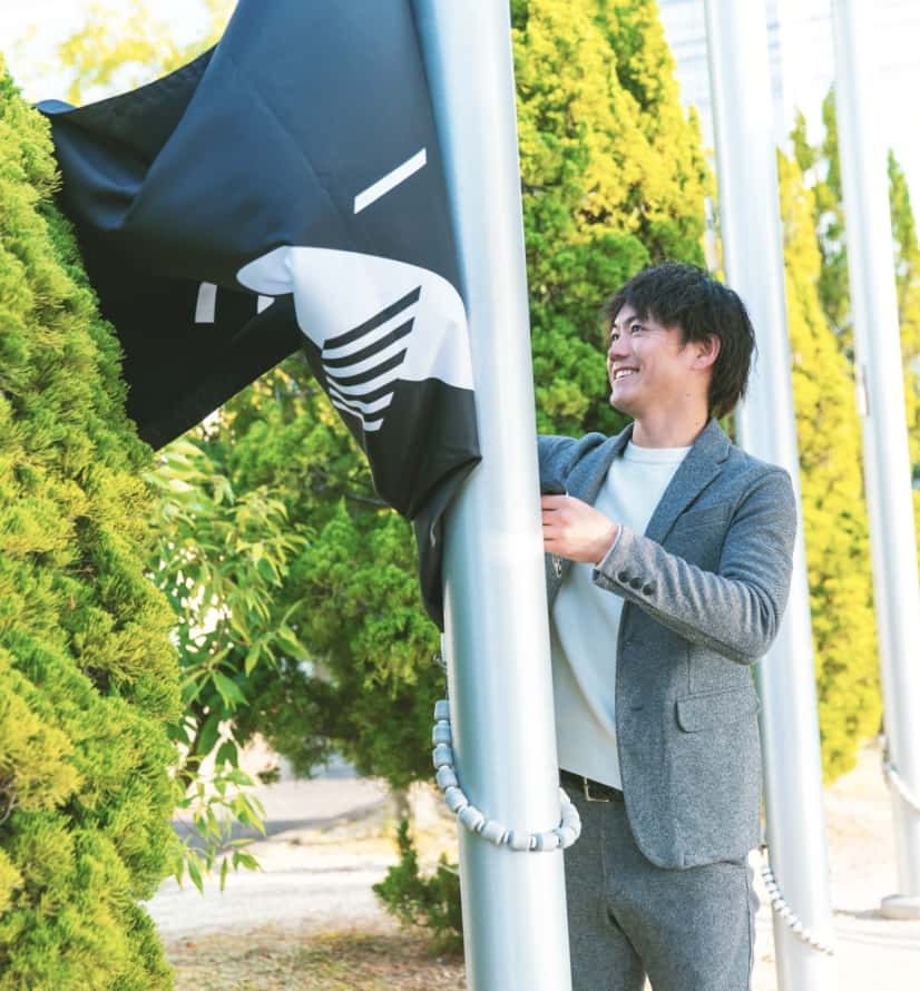 株式会社レニアスのロゴが描かれた大きな黒い旗を持って笑顔を浮かべている、スーツを着た男性社員の写真