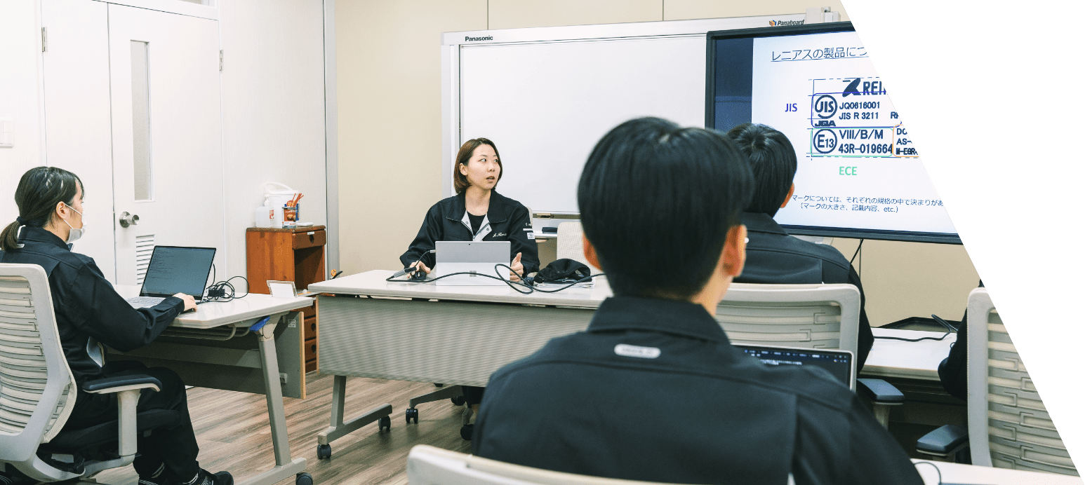 教室のような部屋で、男女スタッフ数名が、前に座っている講師のスタッフの話を聞いている写真