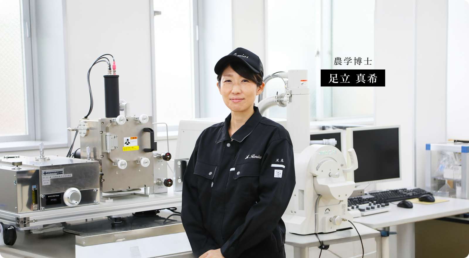 農学博士 足立真希が黒い作業着を来て、機械を背にして立っている写真