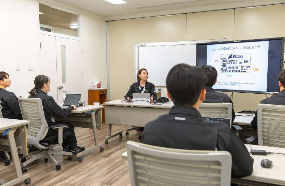 教室のような部屋で、男女スタッフ数名が、前に座っている講師のスタッフの話を聞いている写真。